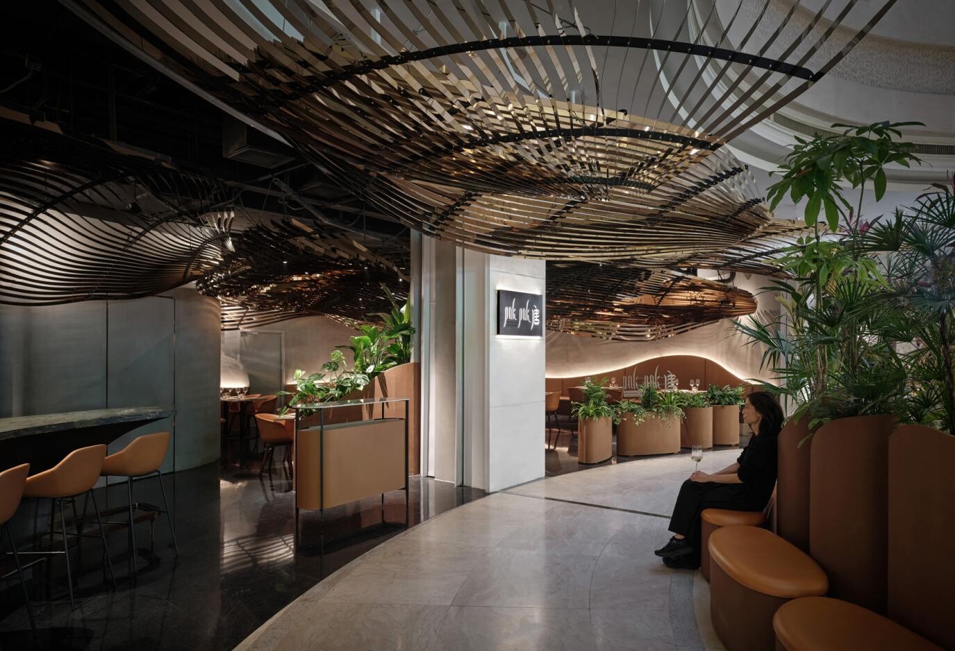 Overall view of the restaurant entrance, where the metal roof installation extends into the dining area and tropical planting with curved seating creates an island-forest atmosphere.